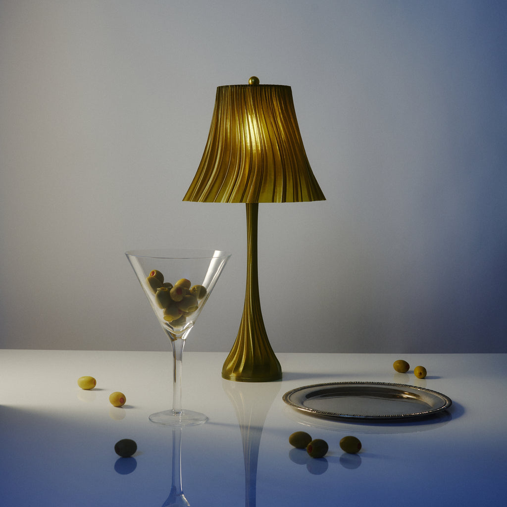 Green lamp, martini glass with olives, and scattered olives on a white surface with a gradient background.