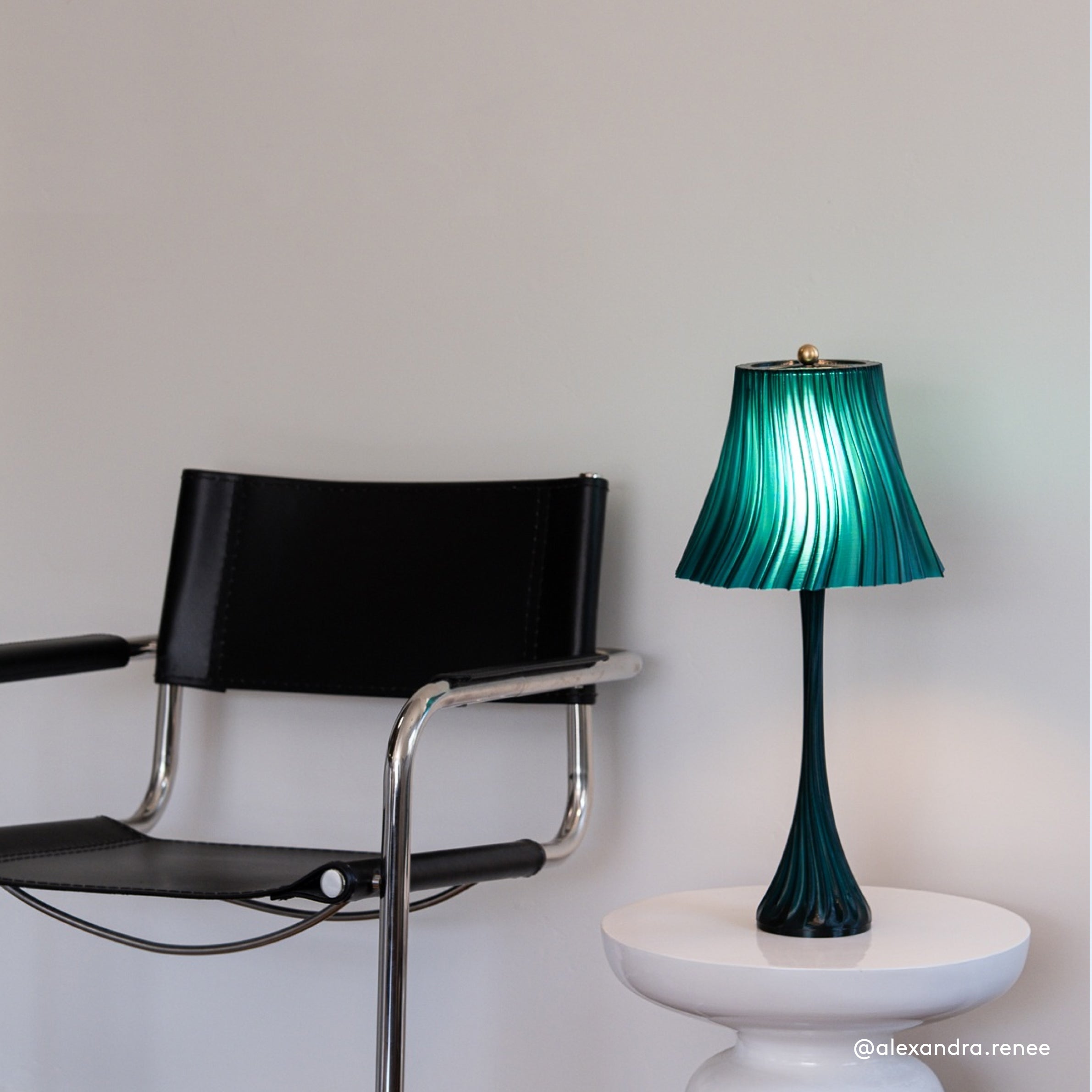 Green lamp on a white table next to a black chair against a light gray wall.