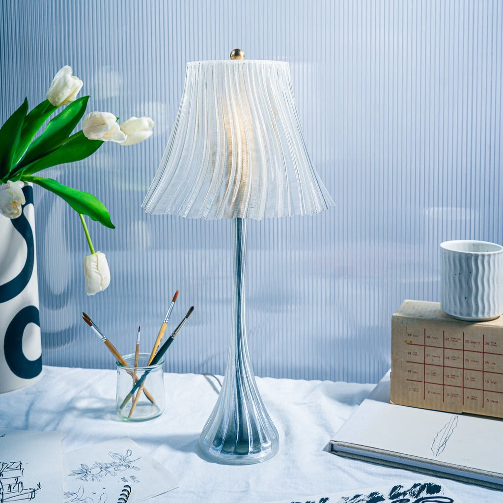 Decorative table lamp with a white shade on a surface with a vase, notebook, and other items.